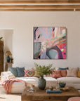 A colorful abstract canvas art print hangs on a white wall in a bright, modern living room with a white sectional sofa, wooden coffee table, and a pink pouf.