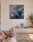 A framed abstract botanical art print with shades of green, purple, and blue hangs on a warm beige wall in a beautifully styled bedroom. The room features a bed with a pink and blue floral throw blanket, a white pleated radiator-style side table, and potted plants.