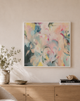 A large square abstract wall art print with soft pastel pinks, blues, yellows, and whites hangs above a light wood console in a bright, modern living room with a cream boucle armchair and natural light.
