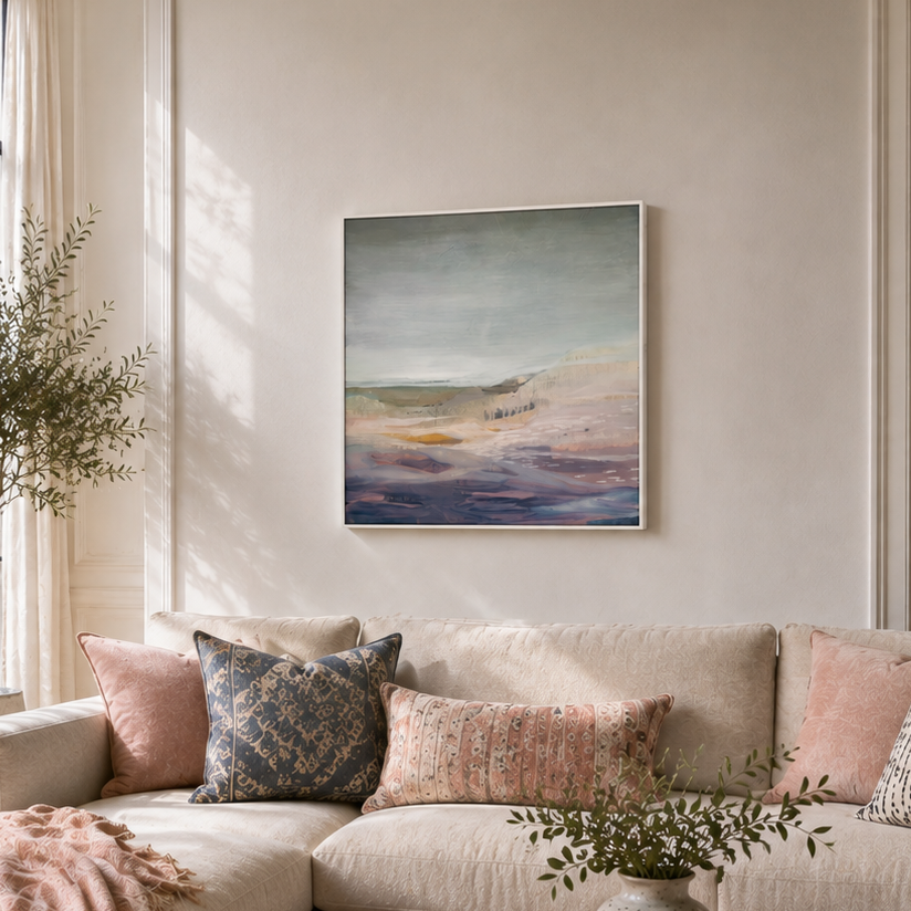 A large beige sectional sofa in a bright, modern living room, styled with pink and patterned throw pillows, a pink blanket, a round wooden coffee table, and a framed abstract landscape painting on the wall.