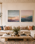 A styled living room featuring two vertical landscape art prints with soft pastel colors hanging above a cream sectional sofa. The room includes a pink armchair, a wooden coffee table, a patterned area rug, a fireplace, and built-in shelves.