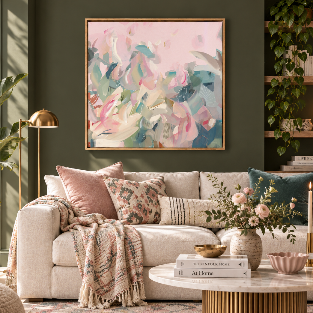 A framed abstract painting with soft pink, teal, and cream brushstrokes hangs on a dark green wall above a beige sofa in a styled living room with pillows, a throw blanket, a coffee table, and plants.