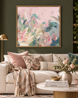 A framed abstract painting with soft pink, teal, and cream brushstrokes hangs on a dark green wall above a beige sofa in a styled living room with pillows, a throw blanket, a coffee table, and plants.