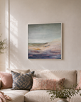 A large beige sectional sofa in a bright, modern living room, styled with pink and patterned throw pillows, a pink blanket, a round wooden coffee table, and a framed abstract landscape painting on the wall.