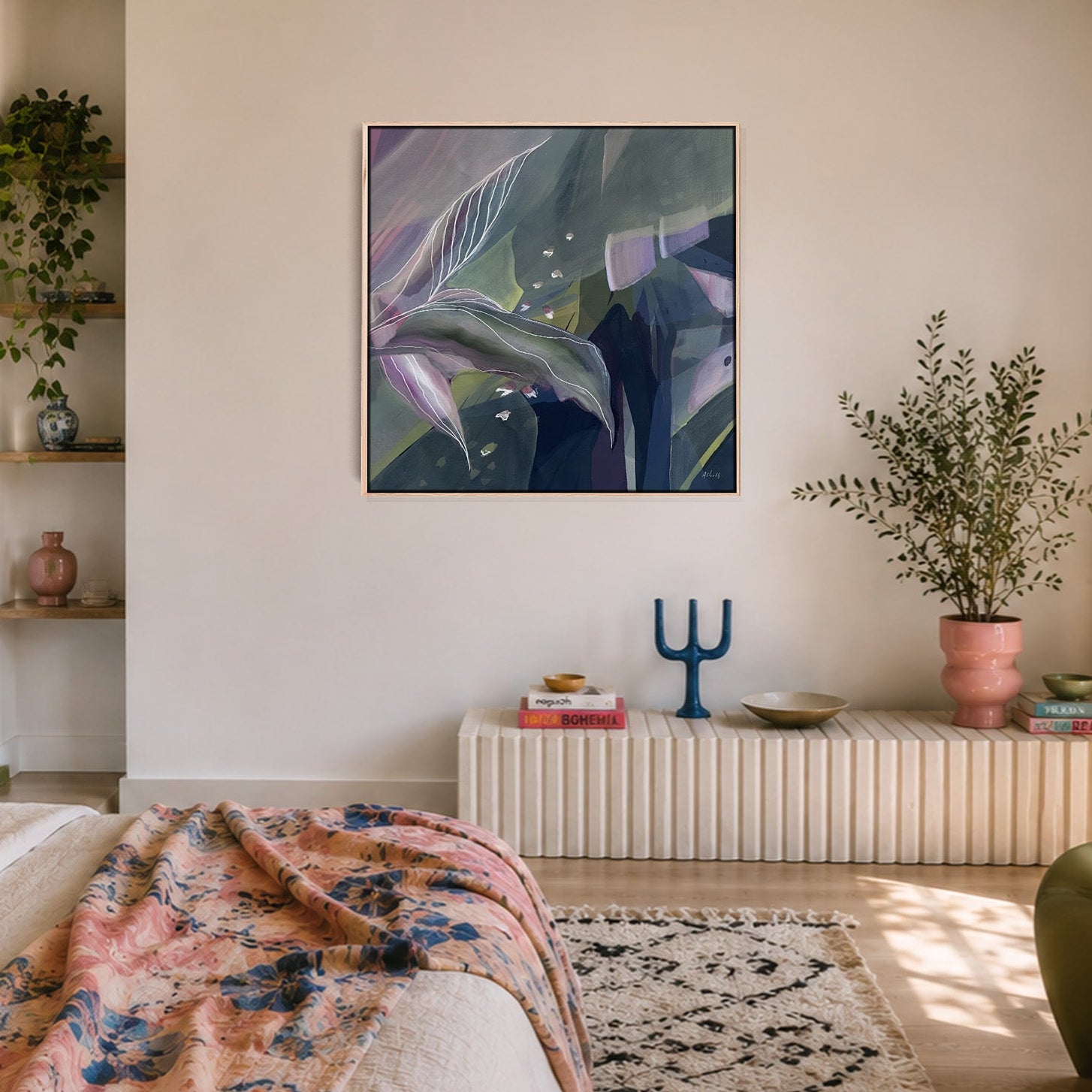 A framed abstract botanical art print with shades of green, purple, and blue hangs on a warm beige wall in a beautifully styled bedroom. The room features a bed with a pink and blue floral throw blanket, a white pleated radiator-style side table, and potted plants.