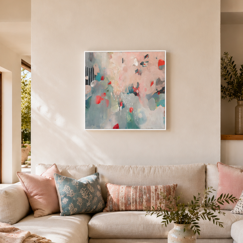 An abstract painting with soft pink, teal, and red brushstrokes hangs on a beige wall above a cream sectional sofa styled with patterned pillows and a throw blanket in a bright, modern living room.